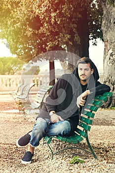 Fashionable cool young man relaxing on a bench in a park with trees
