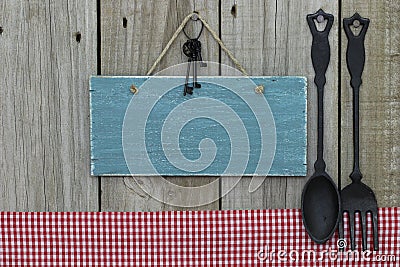 Antique blank blue sign with iron keys, gingham tablecloth and cast iron spoon and fork hanging on wooden background