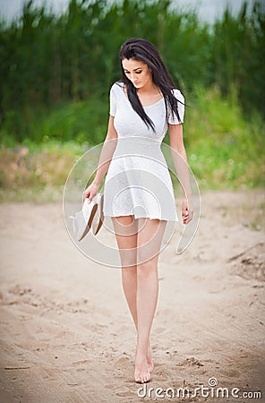 Attractive brunette girl with short white dress strolling barefoot on the countryside road. Young beautiful woman walking