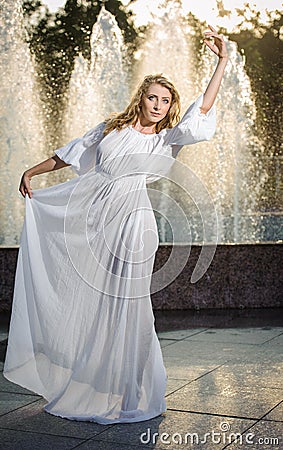 Attractive girl in white long dress sitting in front of a fountain in the summer hottest day