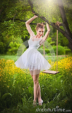Attractive girl in white short dress posing near a tree swing with a flowery meadow in background. Blonde young woman