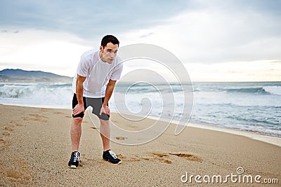 Attractive male runner dressed in the white t-shirt rest standing on the beach looking away
