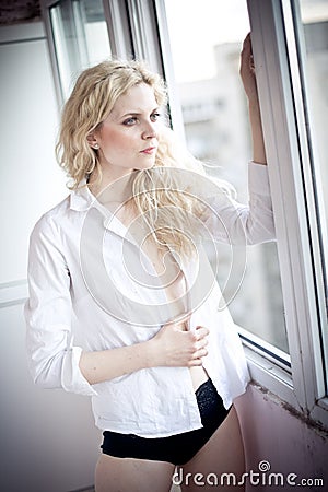 Attractive sexy blonde with white shirt looking on the window in daylight. Portrait of sensual long fair hair woman wearing blouse