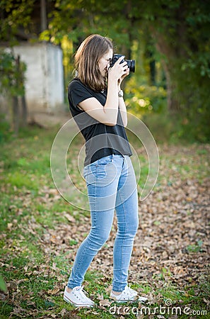Attractive young girl taking pictures outdoors. Cute teenage girl in blue jeans and black t-shirt taking photos in autumnal park.