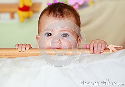 A baby in a cot playing peek-a-boo and sucking the cot rails