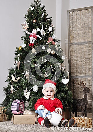 Baby in Santa costume sit near decorating Christmas tree with toy