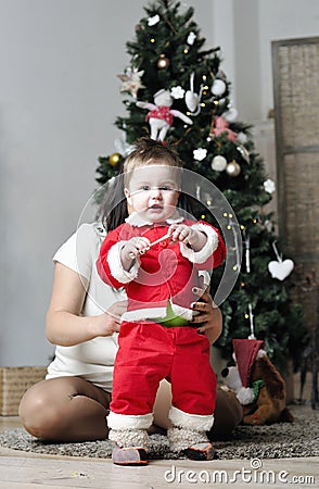 Baby in Santa costume standing with mother on decorating Christmas tree