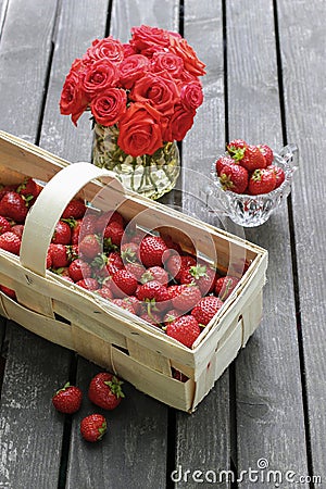 Basket of strawberries on wooden table. Bouquet of red roses in