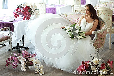 Beautiful bride in a magnificent white wedding dress of tulle with a corset sitting on the sofa with bouquet lily and orchid