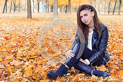 Beautiful girl in rock style with bright makeup in white jacket and black pants and boots in a leather jacket sitting in the Park
