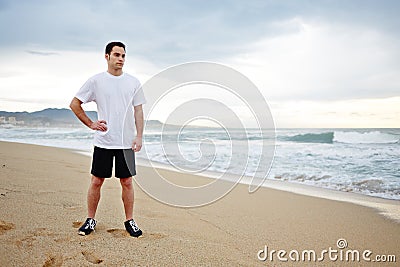 Beautiful male runner dressed in the white t-shirt resting after intensive morning jogging standing on the beach