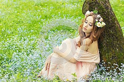 Beautiful sexy girl with red hair with flowers in her hair sitting near a tree in a pink dress in the meadow with blue flowers