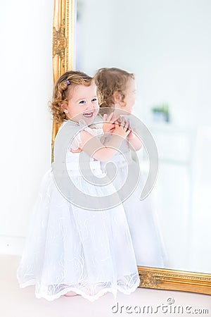 Beautiful toddler girl in white dress next to big mirror