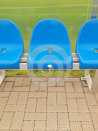 Blue plastic seats on outdoor stadium players bench, chairs with new paint below transparent bended plastic roof.