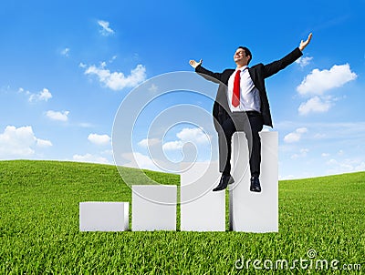 Business Man Outdoors Sitting on a Bar Graph with Arms Raised