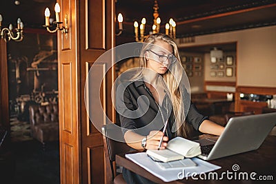 Business woman in glasses indoor with coffee and laptop taking notes in restaurant