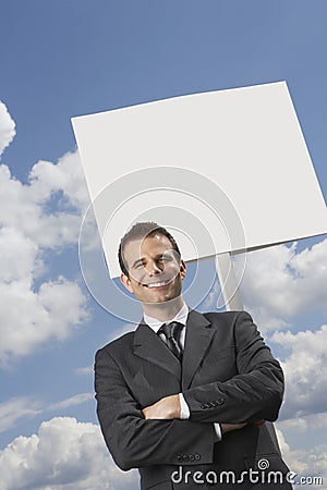 Businessman with arms crossed standing by blank sign against cloudy sky