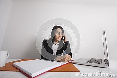 Businesswomen using cell phone while writing notes from laptop at office desk