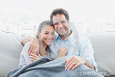 Cheerful couple relaxing on their sofa smiling at camera under blanket