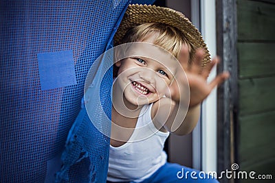 Cheerful kid with straw hat leaning out a window, smiling and waving