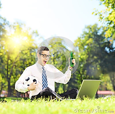 Cheerful man seated on a green grass watching football on a lapt