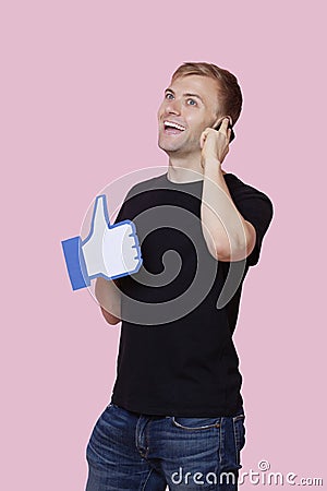 Cheerful young man using cell phone while holding fake like button over pink background