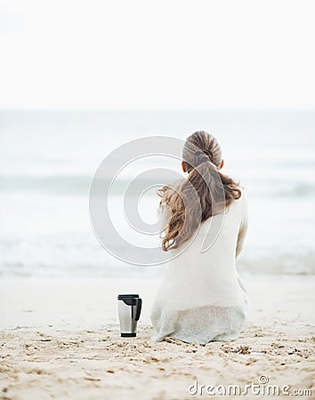Cup of hot beverage near young woman in sweater sitting on beach