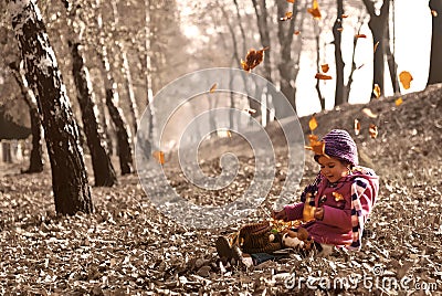 Cute girl sitting on fallen autumn leaves while leafs falling and playing with dolls