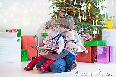 Cute happy boy reading to his toddler sister and newborn baby brother in a dark room with Christmas tree