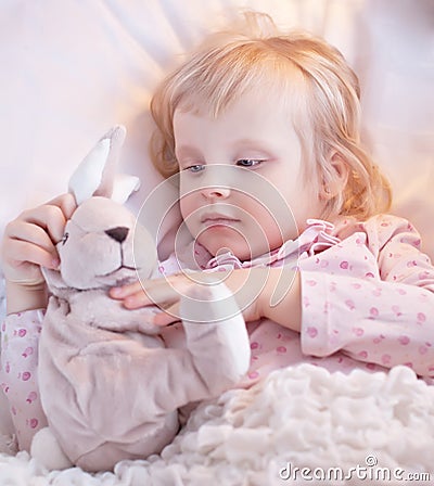 Cute little blond girl play with toy rabbit in bed. Looking ill.