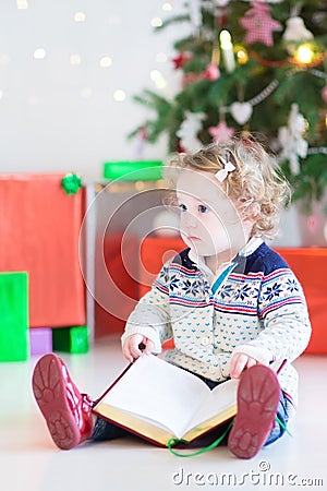 Cute toddler girl reading a book under a Christmas tree