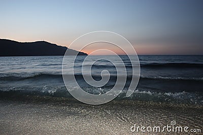 Dark blue sea waves over light sand with mountain and sunset on horizon