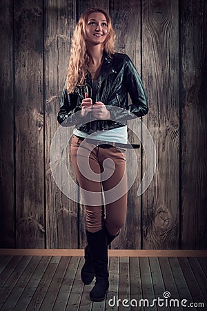 Fashion model with curly hair dressed in black jacket, denim pants and tall boots over wooden wall background