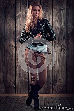 Fashion model with curly hair dressed in black jacket, denim pants and tall boots over wooden wall background