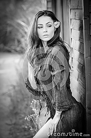 Fashion portrait of sexy brunette in black blouse leaning on wooden cabin wall. Sensual attractive woman with a flower in hair