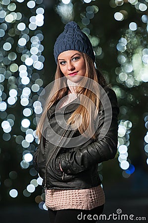 Fashionable lady wearing cap and black jacket outdoor in xmas scenery with blue lights in background. Portrait of young woman