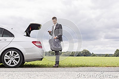Full length of young businessman unloading suitcase from broken down car at countryside