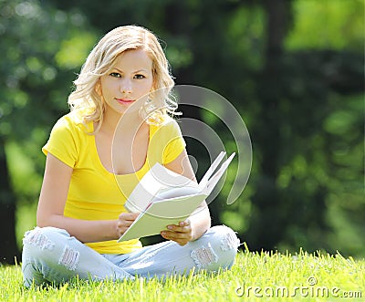 Girl reading the book. Blonde beautiful young woman with book sitting on the grass. Outdoor. Sunny day. Looking at the camera
