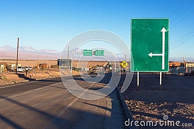 A green road sign stands next to a road in the atacama desert with an arrow going up and one to the right