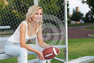 Happy blonde girl with american football. Smiling cheerful beautiful young woman sitting on the bench. Outdoors. Fan of football