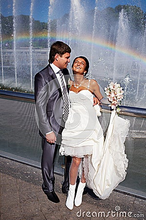 Happy bride and groom near fountain with rainbow