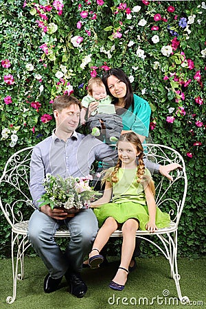 Happy family of four sit on white bench with bunch of flowers