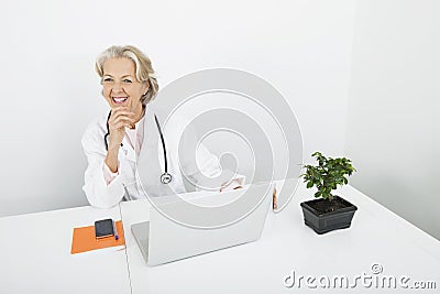 Happy senior female doctor with laptop at desk in clinic