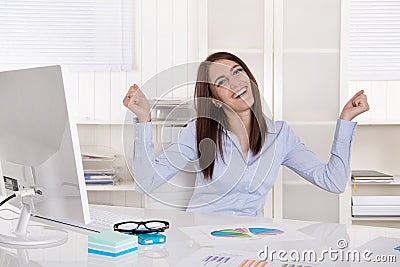 Happy successful young business woman with arms up at desk.