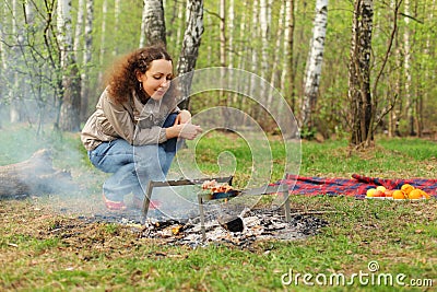 Happy woman sits near campfire with grill