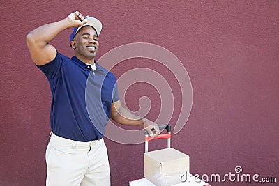 Happy young African American delivery man standing with handtruck over colored background