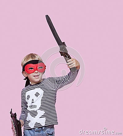 Happy young boy wearing eye mask holding sword and shield over pink background