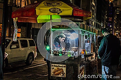 Hot dog vendor serving a customer from his cart on a nighttime Manhattan sidewalk