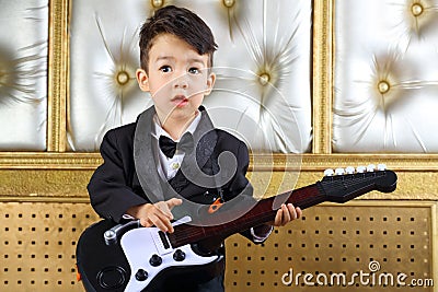 A little boy in black tuxedo stands with guitar