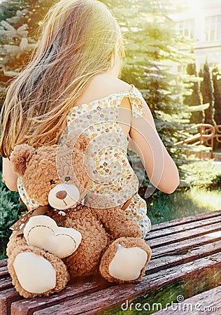 Little girl sitting back with teddy bear on the bench in sun bea